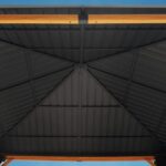 Enjoy the view from beneath the 10 ft x 10 ft Wood Grain Outdoor Patio Gazebo with Metal Roof, featuring a geometric-patterned hardtop canopy supported by elegant wooden-style beams, set against a clear blue sky.